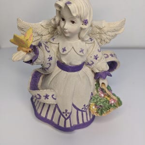 Puede incluir: Figura de ángel de cerámica sentada, vestida con un vestido blanco con detalles morados, sosteniendo una mariposa amarilla. El ángel tiene alas emplumadas y una cesta de flores. La figura mide aproximadamente 15 cm de altura.
