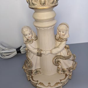 Puede incluir: Lámpara de mesa color crema con una base decorativa con dos figuras de niñas tomadas de la mano. La lámpara tiene detalles dorados y un diseño de columna detallado. La lámpara está enchufada y lista para usar.