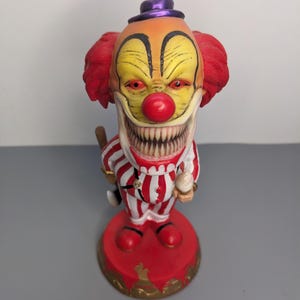 Puede incluir: Una figura de payaso con cara amarilla, ojos rojos y una boca ancha llena de dientes afilados. El payaso viste un traje a rayas rojas y blancas, un pequeño sombrero morado y tiene el pelo rojo. Sostiene un pequeño objeto blanco sobre una base roja.