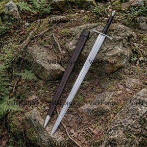 May include: A long, silver sword with a black handle and crossguard, resting on a mossy rock. A brown leather scabbard lies beside it. The sword's blade is polished, reflecting the light. The overall theme is medieval weaponry.