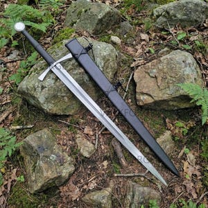 May include: A long, silver sword with a black handle and crossguard, resting on a bed of rocks and foliage. The sword is accompanied by a black sheath. The blade is approximately 36 inches long.