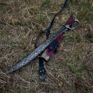 Könnte beinhalten: Ein geschwungenes, verziertes Schwert mit gemusterter Klinge und dekorativem Griff liegt auf einer weinroten Scheide. Die Scheide hat ein detailliertes metallisches Design und wird von einem schwarzen Lederriemen gehalten. Das Schwert liegt auf trockenem Gras.