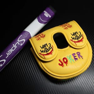 May include: A yellow golf putter head cover with the word "JOKER" in red, green, and purple embroidery. The cover has the phrase "WHY SO SERIOUS?" and "HA HA HA" embroidered in red. A purple and white golf club is next to the cover.
