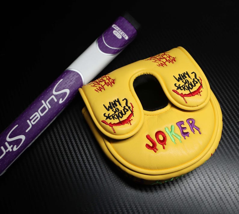 May include: A yellow golf putter head cover with the word "JOKER" in red, green, and purple embroidery. The cover has the phrase "WHY SO SERIOUS?" and "HA HA HA" embroidered in red. A purple and white golf club is next to the cover.