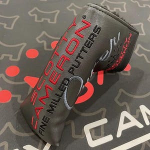 Puede incluir: Funda de putter de golf negra con texto bordado en rojo que dice "SCOTTY CAMERON FINE MILLED PUTTERS". La funda tiene un diseño gris y negro en la superficie. El interior de la funda es suave y negro.