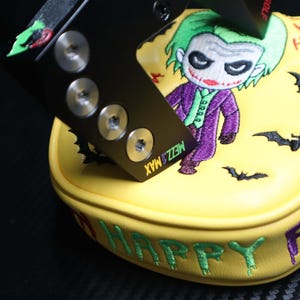 May include: Yellow golf club head cover with an embroidered cartoon Joker character in a purple suit and green hair. The cover has the words "HAPPY FR" and "HA HA" embroidered in green and red. A black metal piece with screws is on top.