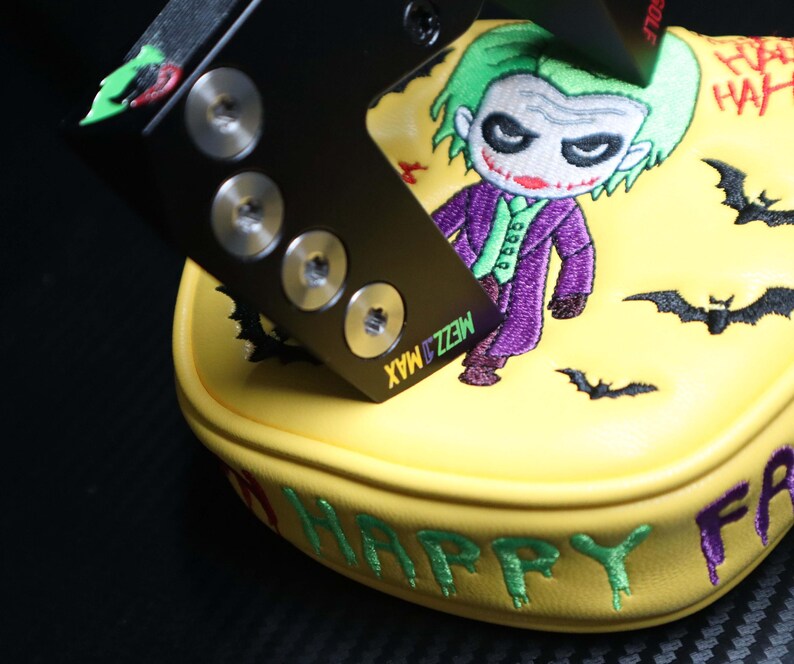 May include: Yellow golf club head cover with an embroidered cartoon Joker character in a purple suit and green hair. The cover has the words "HAPPY FR" and "HA HA" embroidered in green and red. A black metal piece with screws is on top.