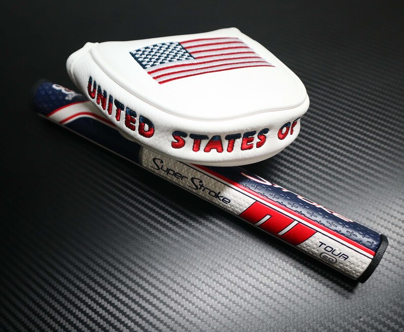 May include: White golf club head cover with an embroidered American flag and the words "UNITED STATES OF" in red. A golf club grip with red, white, and blue stripes and the text "Super Stroke" is also visible.