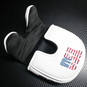 May include: A white and black golf putter cover with the word "USA" embroidered in red, white, and blue. The cover has a padded interior and a magnetic closure. The putter cover is designed to protect the golf club head.
