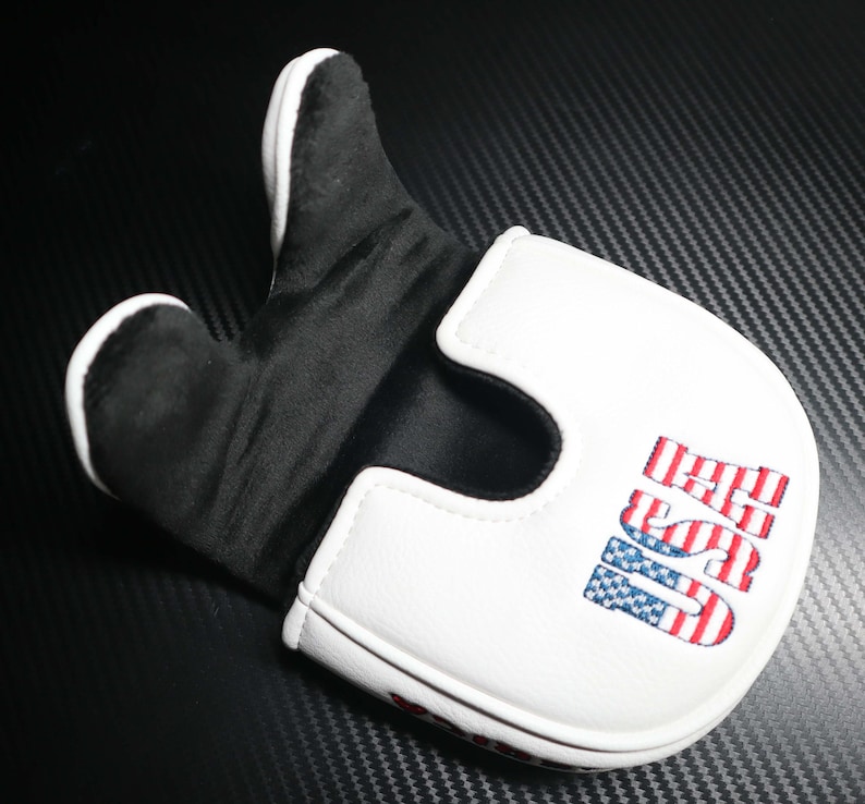 May include: A white and black golf putter cover with the word "USA" embroidered in red, white, and blue. The cover has a padded interior and a magnetic closure. The putter cover is designed to protect the golf club head.