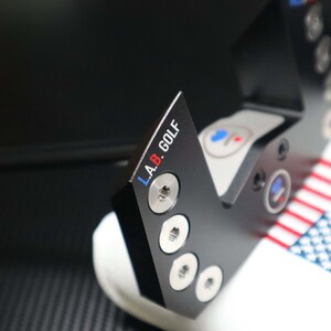 May include: Black golf putter head with the text "L.A.B. GOLF" in white, blue, and red. The putter head has a series of silver screws and circular accents. The putter is resting on a white surface with a red, white, and blue striped pattern.