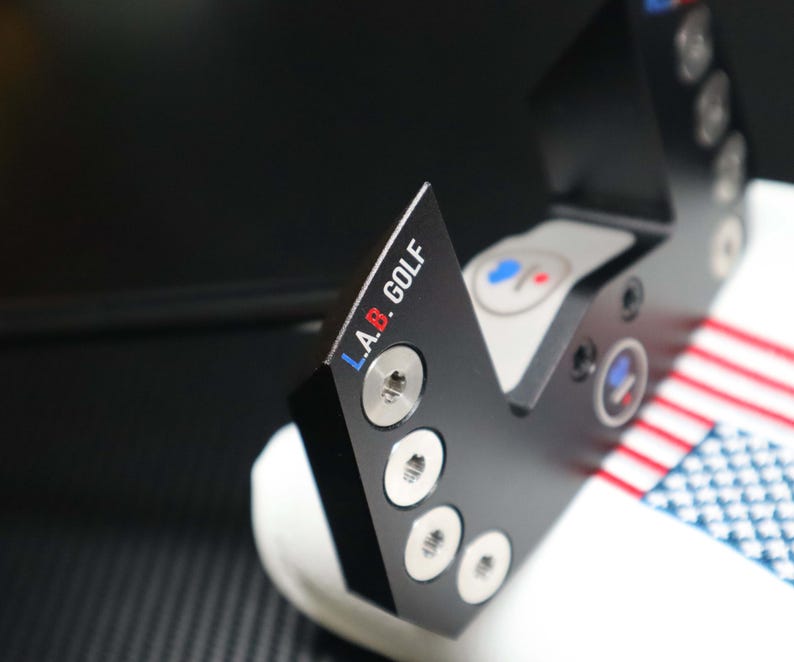 May include: Black golf putter head with the text "L.A.B. GOLF" in white, blue, and red. The putter head has a series of silver screws and circular accents. The putter is resting on a white surface with a red, white, and blue striped pattern.