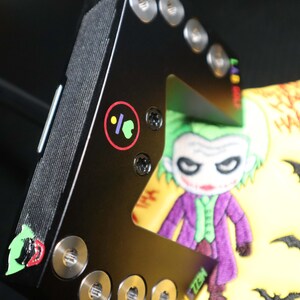 May include: Black metal object featuring a cartoon Joker design. The Joker is in a purple suit, green hair, and white face. The object has silver screws and a colourful logo.