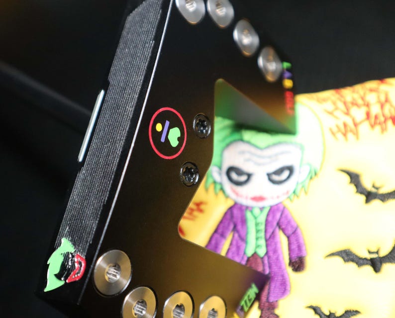 May include: Black metal object featuring a cartoon Joker design. The Joker is in a purple suit, green hair, and white face. The object has silver screws and a colourful logo.