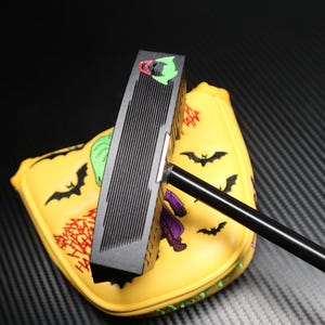 May include: A black golf putter with a yellow head cover featuring a cartoon design. The head cover has a bat and character design. The putter has a black shaft and a black head with vertical grooves. The head cover has a red and green design.