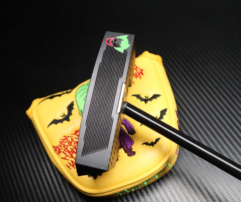 May include: A black golf putter with a yellow head cover featuring a cartoon design. The head cover has a bat and character design. The putter has a black shaft and a black head with vertical grooves. The head cover has a red and green design.