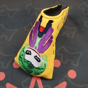 Puede incluir: Funda amarilla para la cabeza del palo de golf con una imagen bordada del Joker. La funda tiene un interior negro y bordados morados, verdes y blancos. La funda está hecha de un material suave.