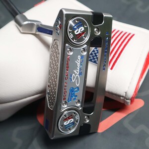 May include: A Scotty Cameron Studio Style golf putter with a silver finish, featuring the text "USA" in blue and red accents. The putter is paired with a white headcover with red and blue stripes, and the text "SQUAREBACK 15".