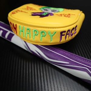 May include: Yellow golf club head cover featuring an embroidered Joker design and the words "ON HAPPY FACE" in green and purple. The golf club handle is purple and white.