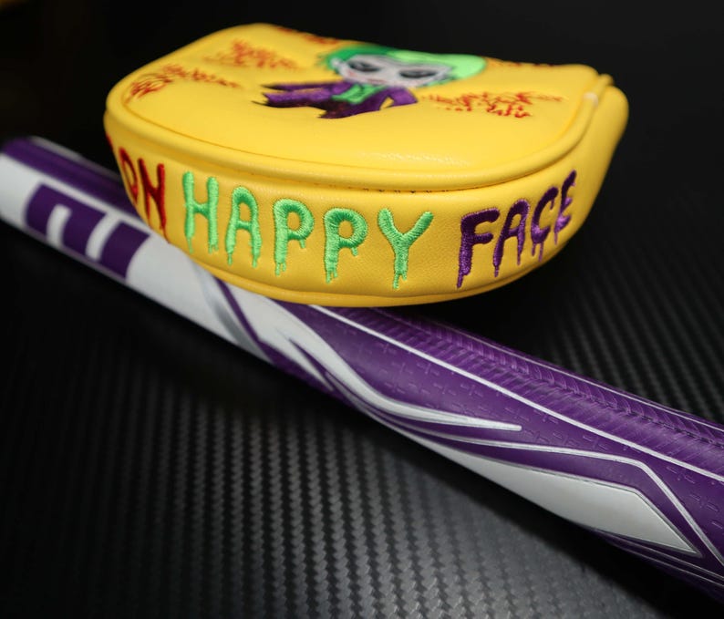 May include: Yellow golf club head cover featuring an embroidered Joker design and the words "ON HAPPY FACE" in green and purple. The golf club handle is purple and white.