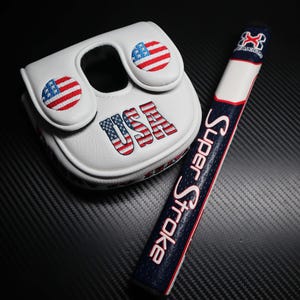 May include: White golf putter cover and grip with patriotic USA theme. The cover features embroidered American flags and the word "USA". The grip is navy blue with the words "Super Stroke" in pink.