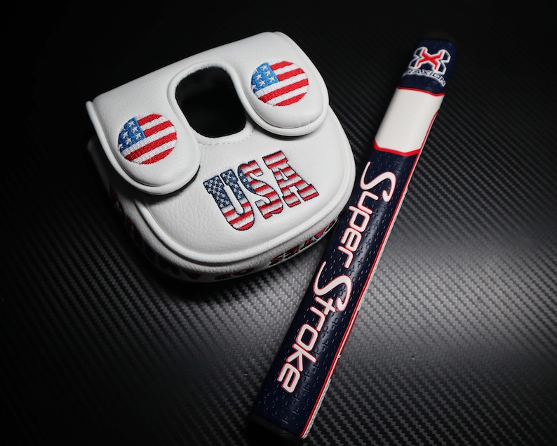 May include: White golf putter cover and grip with patriotic USA theme. The cover features embroidered American flags and the word "USA". The grip is navy blue with the words "Super Stroke" in pink.