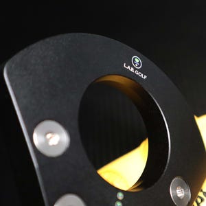 May include: Close-up of a black golf club putter head with a circular opening. The putter has the text "LAB GOLF" and a logo on the top. Silver screws are visible on the putter head. The background is black.