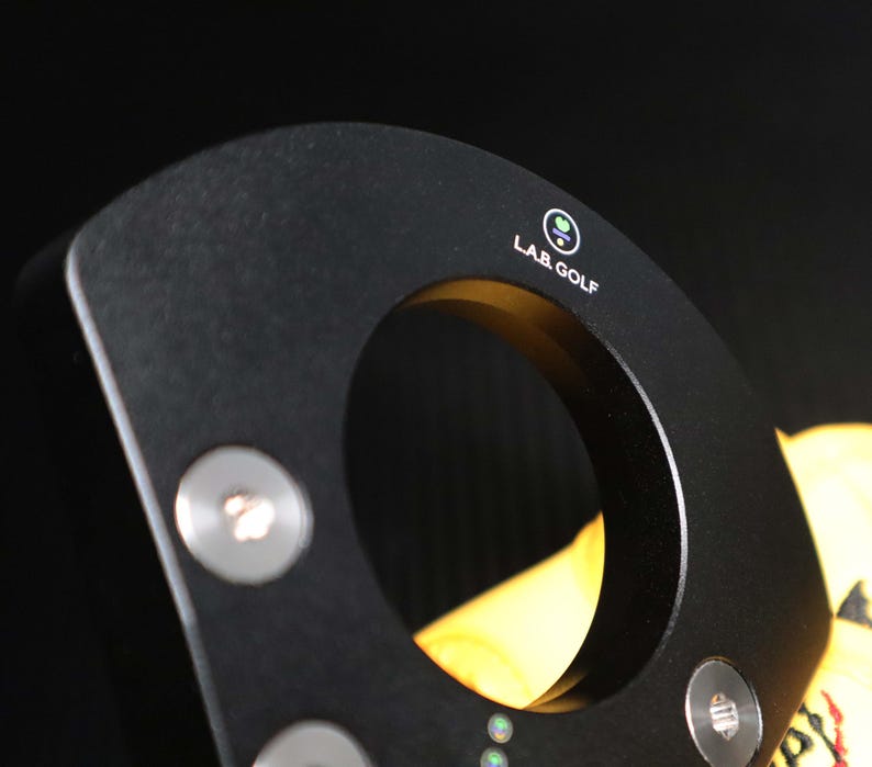May include: Close-up of a black golf club putter head with a circular opening. The putter has the text "LAB GOLF" and a logo on the top. Silver screws are visible on the putter head. The background is black.