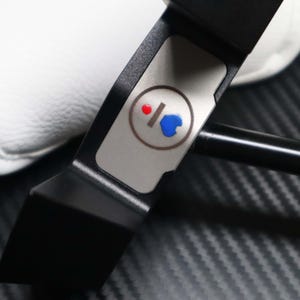 May include: A black golf putter with a silver accent featuring a red and blue design. The putter has a sleek, modern design and a white golf club cover is visible in the background.