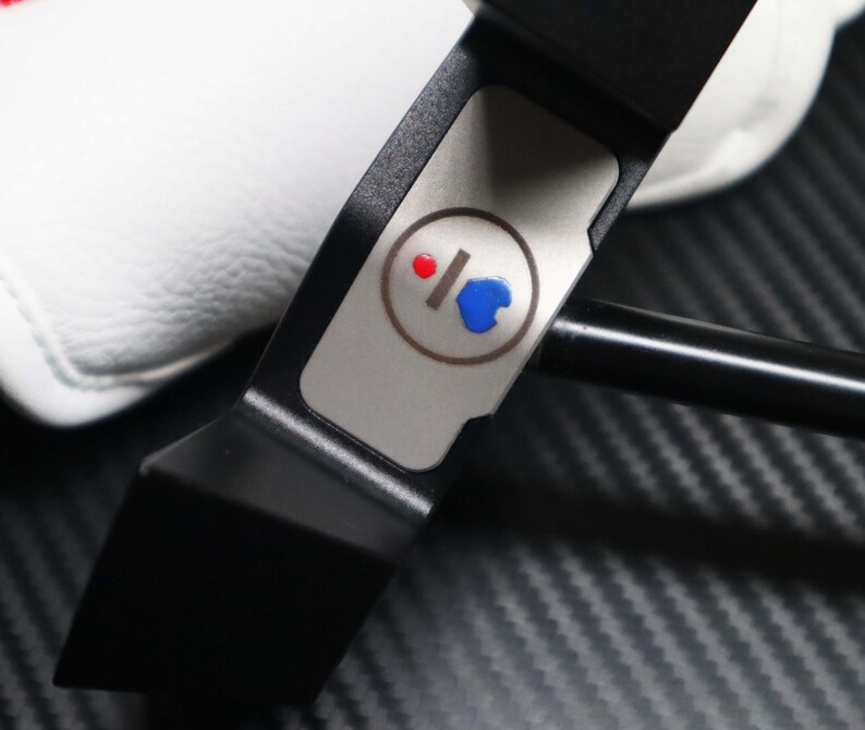 May include: A black golf putter with a silver accent featuring a red and blue design. The putter has a sleek, modern design and a white golf club cover is visible in the background.