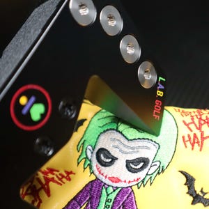 May include: Black golf club head cover featuring an embroidered Joker design. The cover is yellow with a green-haired Joker, purple suit, and the words "HA HA HA". The club has the text "L.A.B. GOLF".