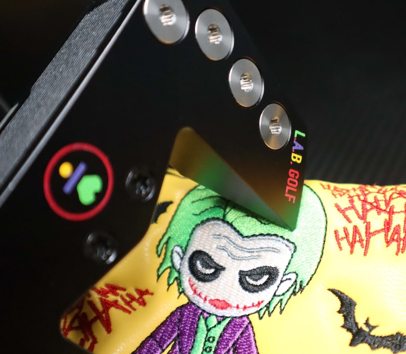 May include: Black golf club head cover featuring an embroidered Joker design. The cover is yellow with a green-haired Joker, purple suit, and the words "HA HA HA". The club has the text "L.A.B. GOLF".