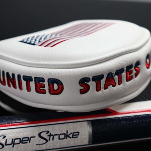 May include: White golf club head cover with the words "UNITED STATES OF" embroidered in red and blue. The top features an embroidered American flag. A golf club grip with the words "Super Stroke" is visible.
