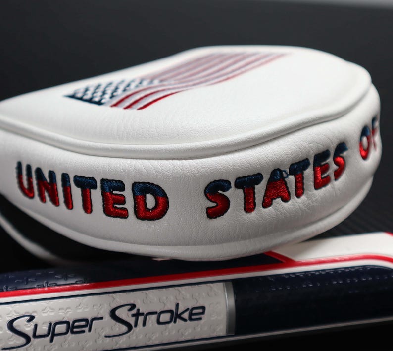 May include: White golf club head cover with the words "UNITED STATES OF" embroidered in red and blue. The top features an embroidered American flag. A golf club grip with the words "Super Stroke" is visible.