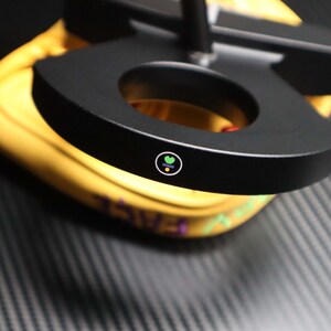 May include: A black, circular object with a hole in the center, possibly a part of a lamp or stand. The object has a small, colorful logo on its surface. The background is yellow and the surface is dark gray.