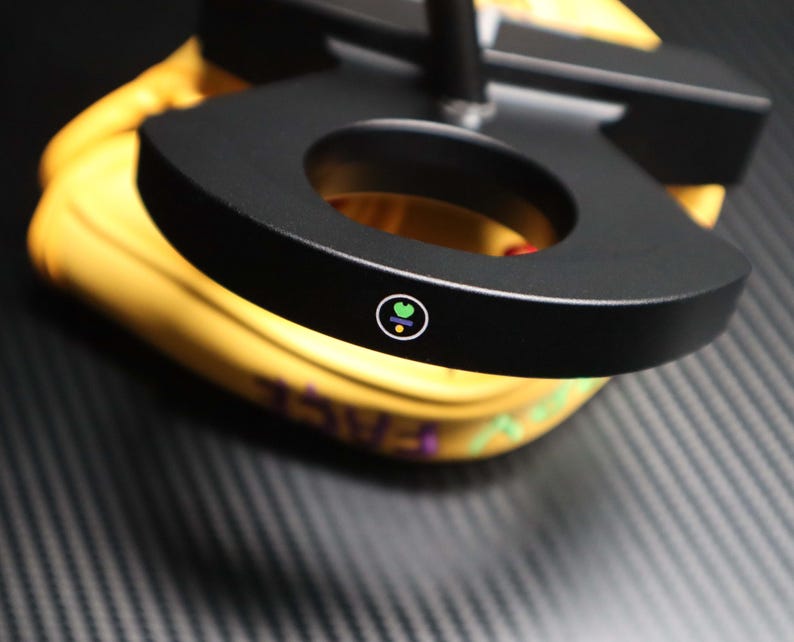 May include: A black, circular object with a hole in the center, possibly a part of a lamp or stand. The object has a small, colorful logo on its surface. The background is yellow and the surface is dark gray.