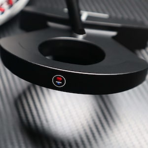 May include: A black golf putter with a circular design and a central opening. The putter features a red and blue circular emblem on the side. The putter is set against a textured, dark gray surface.