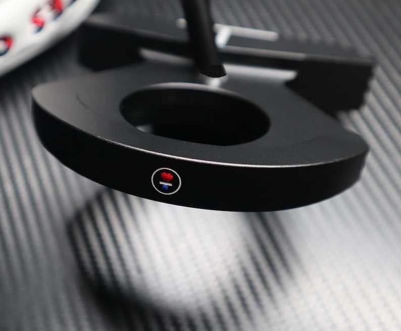 May include: A black golf putter with a circular design and a central opening. The putter features a red and blue circular emblem on the side. The putter is set against a textured, dark gray surface.
