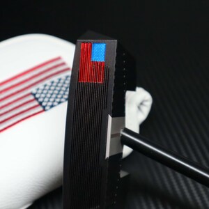 May include: A black golf putter with a red and blue American flag design on the head. The putter has a black shaft and is next to a white golf club cover with an American flag design.