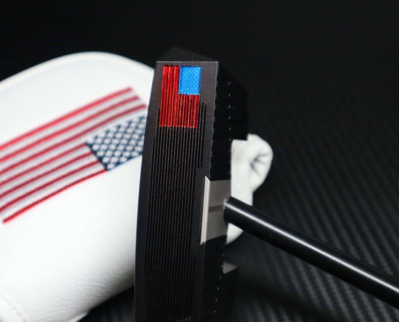 May include: A black golf putter with a red and blue American flag design on the head. The putter has a black shaft and is next to a white golf club cover with an American flag design.