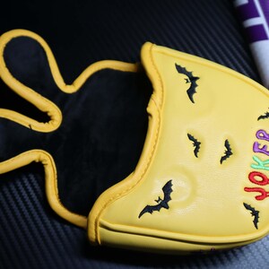May include: Yellow golf club head cover with a black interior and a yellow exterior. The cover features embroidered black bats and the word "JOKER" in green, red, and purple. The cover has a unique design.