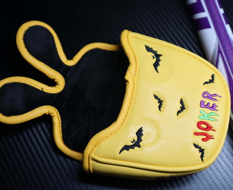 May include: Yellow golf club head cover with a black interior and a yellow exterior. The cover features embroidered black bats and the word "JOKER" in green, red, and purple. The cover has a unique design.