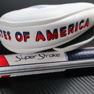 May include: A white golf club head cover with the words "STATES OF AMERICA" embroidered in red and blue. The cover is resting on a golf club grip with the text "Super Stroke". The design incorporates the colors of the American flag.