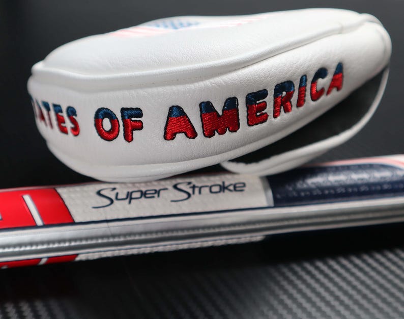 May include: A white golf club head cover with the words "STATES OF AMERICA" embroidered in red and blue. The cover is resting on a golf club grip with the text "Super Stroke". The design incorporates the colors of the American flag.