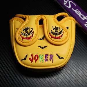 May include: Yellow golf club head cover with the word "JOKER" in colourful embroidery. The cover features the phrase "WHY SO SERIOUS?" and bat silhouettes. A purple and white golf club is visible in the background.
