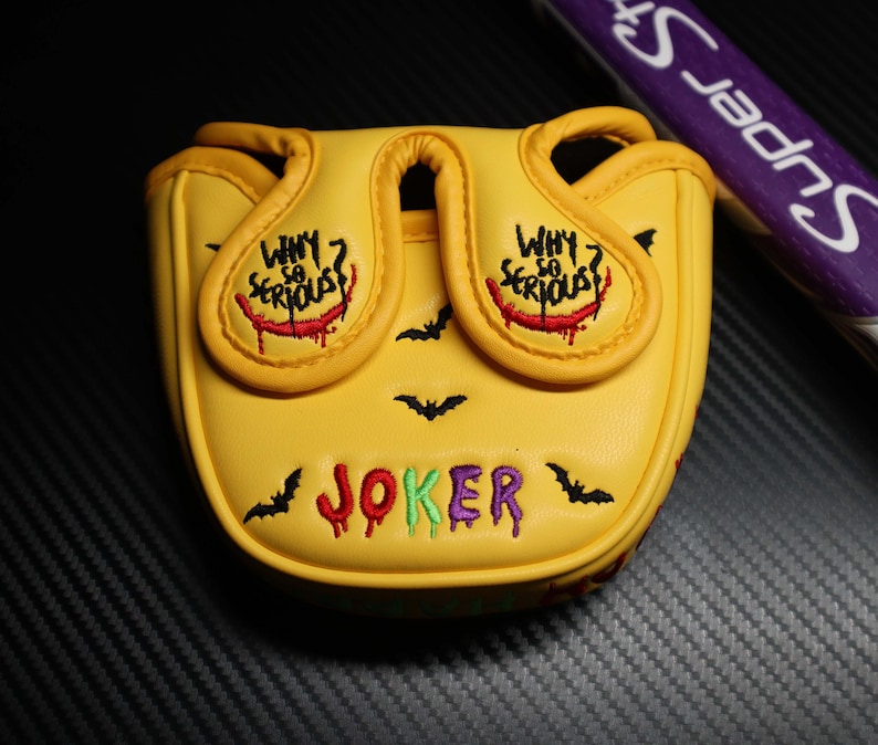 May include: Yellow golf club head cover with the word "JOKER" in colourful embroidery. The cover features the phrase "WHY SO SERIOUS?" and bat silhouettes. A purple and white golf club is visible in the background.