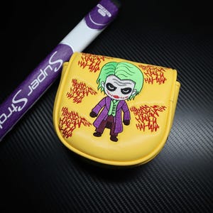 May include: Yellow golf club head cover featuring an embroidered cartoon image of the Joker with green hair and purple suit. The cover has red embroidered "HA HA HA" text around the Joker. A purple and white golf club is in the background.