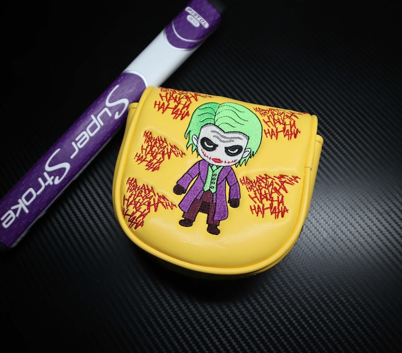 May include: Yellow golf club head cover featuring an embroidered cartoon image of the Joker with green hair and purple suit. The cover has red embroidered "HA HA HA" text around the Joker. A purple and white golf club is in the background.