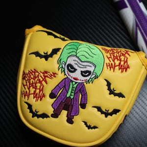 May include: Yellow golf club head cover with an embroidered cartoon Joker design. The cover features green hair, a purple coat, and red lipstick. Black bat silhouettes and red "HA HA HA" text are also present.