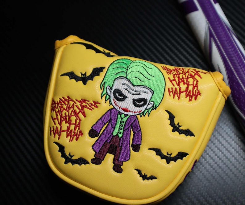 May include: Yellow golf club head cover with an embroidered cartoon Joker design. The cover features green hair, a purple coat, and red lipstick. Black bat silhouettes and red "HA HA HA" text are also present.